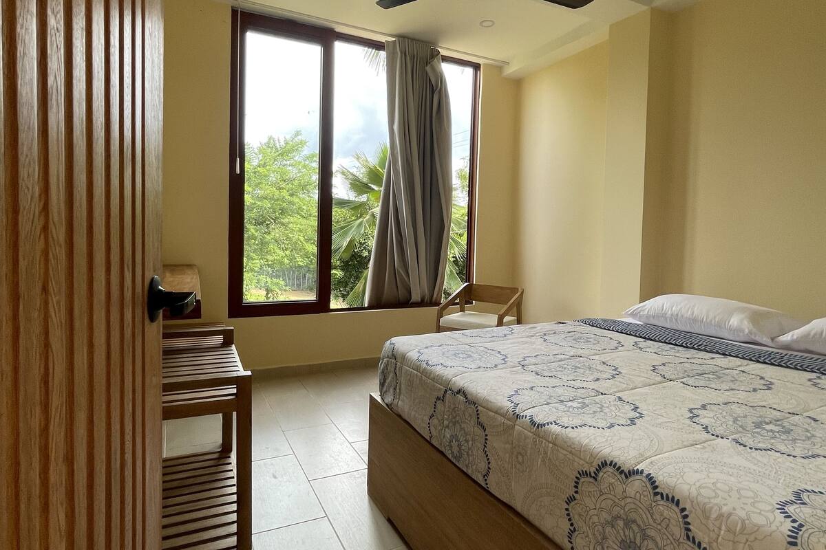 A bedroom with a bed, a ceiling fan, a window with curtains, a chair, and a wooden door.