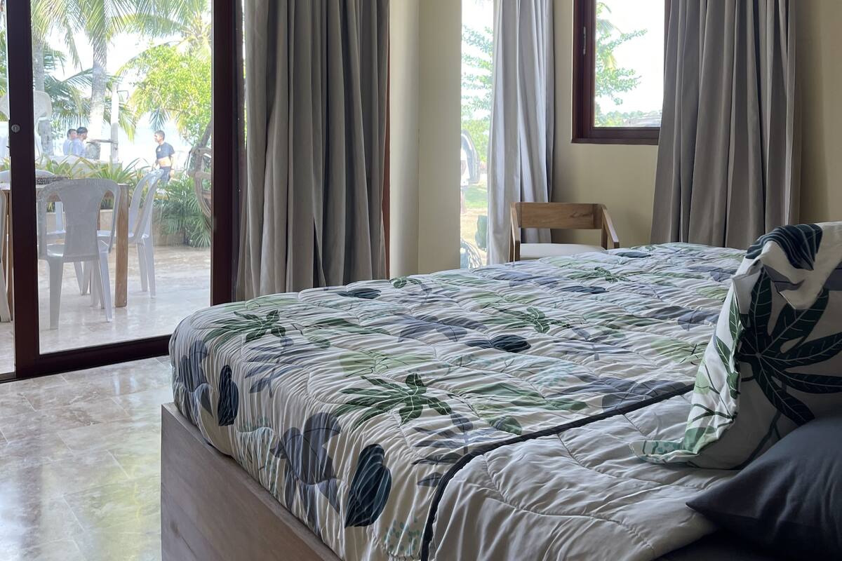 A bedroom with a bed, a ceiling fan, and a view of an outdoor area with chairs and palm trees.
