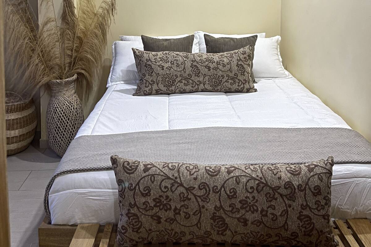 A bedroom with a bed, pillows, a ceiling fan, an air conditioner, and decorative plants.