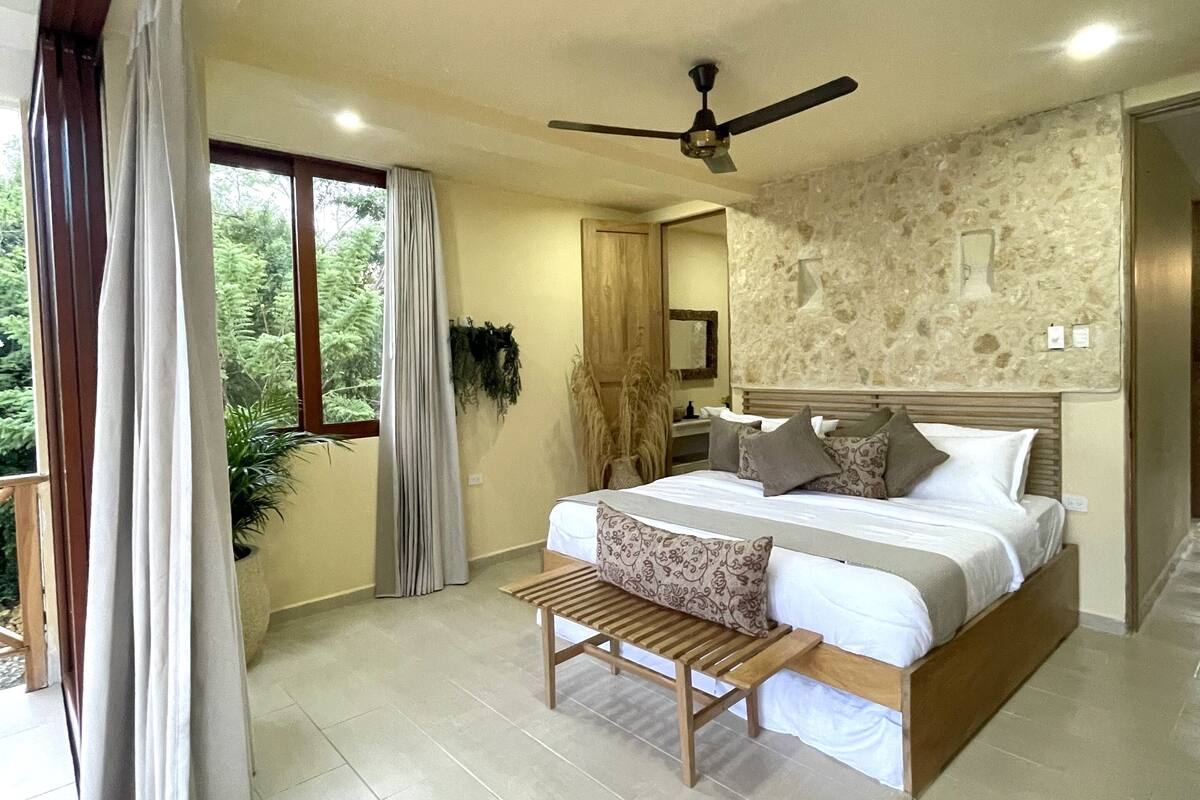 A spacious bedroom with a large bed, a ceiling fan, and a view of greenery outside.