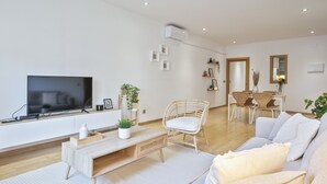 Una sala de estar moderna con un televisor de pantalla plana, una mesa de centro de madera, una silla de mimbre, un sofá y una zona de comedor con mesa y sillas.