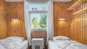 A room with wooden walls, two beds, a window with white curtains, and a small heater.