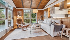 A modern living room with a wooden ceiling, large windows, a white sofa, a leather armchair, and a round coffee table.