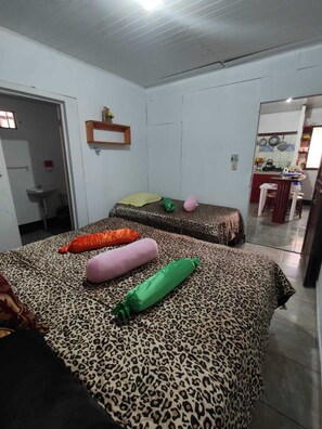 Ein Zimmer mit zwei Betten, jedes mit leopardenmuster-Bettwäsche und vielen Kissen. An der Wand befindet sich eine kleine Ablagefläche mit einer Pflanze, und im Hintergrund ist eine Küchenzeile erkennbar.