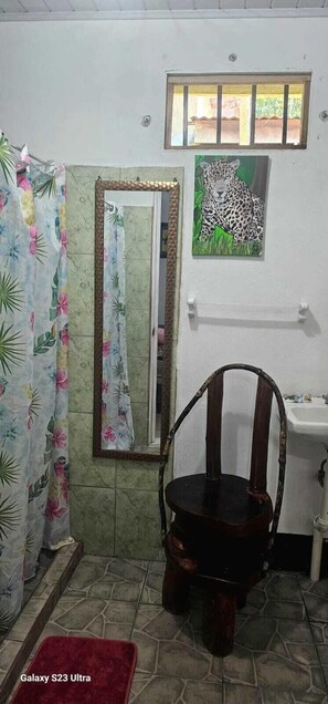 Ein Badezimmer mit Duschvorhang, Spiegel, einem Leopardenbild, einem Holzhocker, Waschbecken und einem Galaxy S23 Ultra Smartphone auf dem Boden.