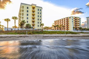 Un hotel frente al mar con varios pisos, una playa de arena en primer plano y palmeras.