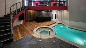 Una piscina interior moderna con un solárium de madera, un jacuzzi y una escalera que conduce a un nivel superior.