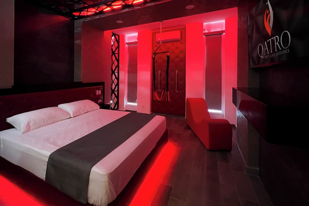 A modern hotel room with a large bed, a red accent wall, and a red-lit reception desk.