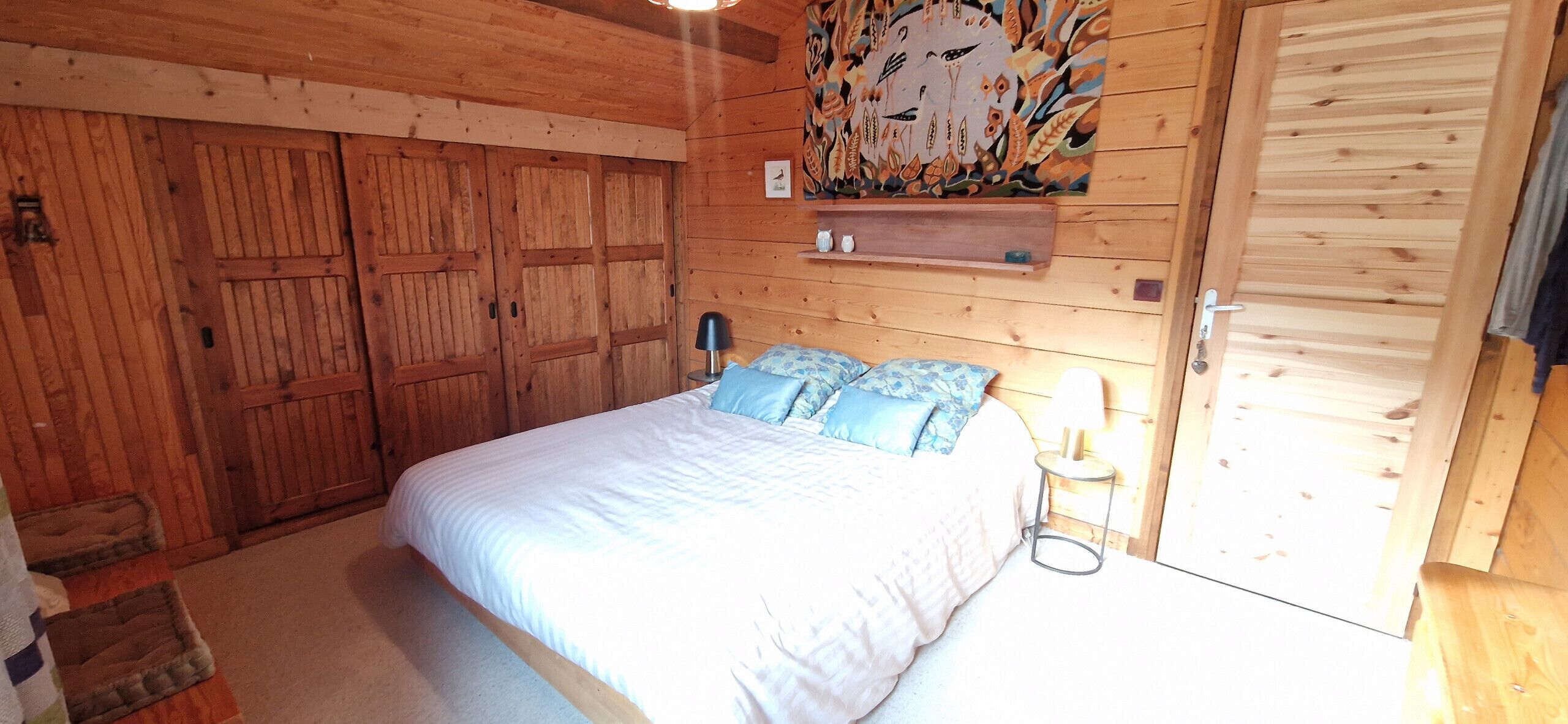A wooden-paneled bedroom with a bed, pillows, a nightstand, and a shelf with decorative items.