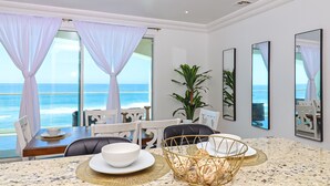 Un comedor con vista al océano, cortinas blancas y una mesa servida con platos y tazones.