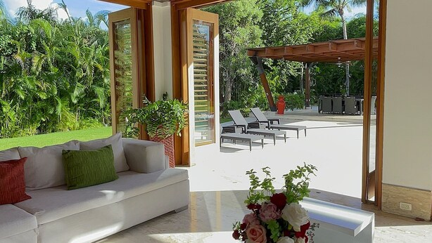 Ein geräumiger Wohnbereich mit einem weißen Sofa, einem Couchtisch mit Blumenvase und Blick auf einen Garten durch große Fenster.