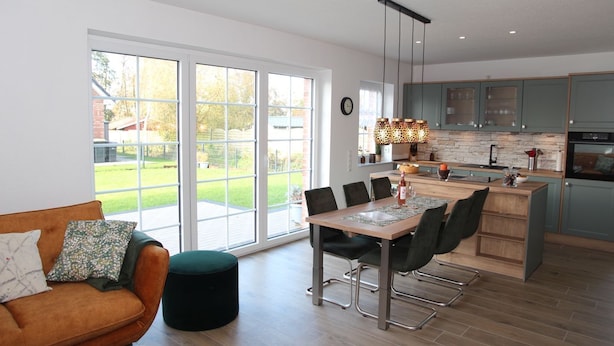 A modern kitchen with large windows, a dining area with a table and chairs, and a cozy living room with a sofa and ottoman.