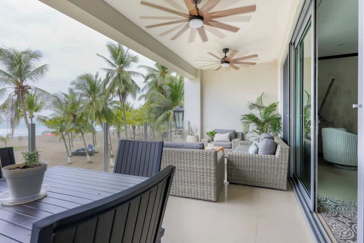 Een balkon met rieten meubilair, een tafel en plafondventilatoren. Palmbomen en een strand zijn op de achtergrond zichtbaar.