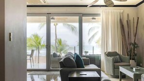 Una sala de estar moderna con un gran ventanal corredizo que da a un balcón con vistas a palmeras y al océano. Hay un sofá, una mesa de centro y una silla de mimbre.