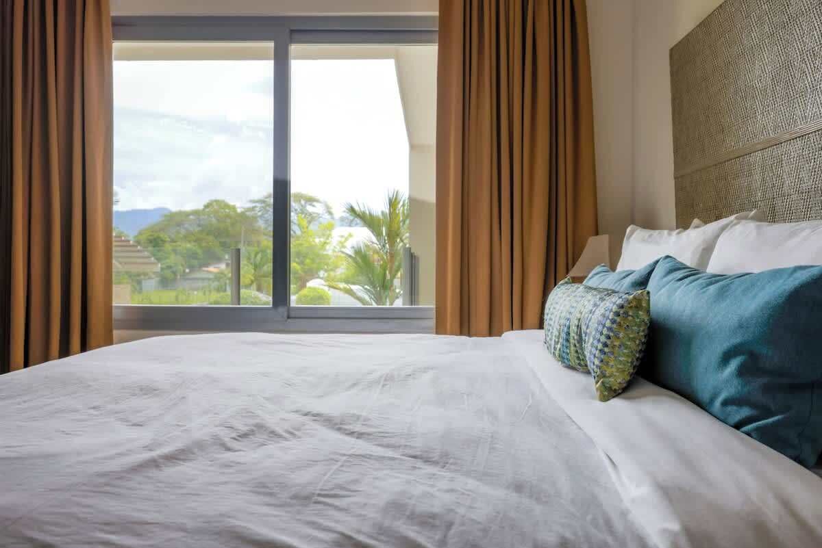 A hotel room with a large bed, a view of greenery and buildings through a sliding door, and a patterned headboard.