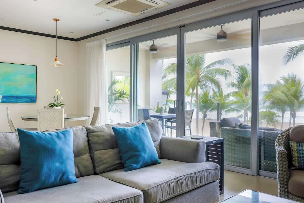 A modern living room with a grey sofa, two blue cushions, a glass coffee table, and a view of palm trees through large sliding glass doors.