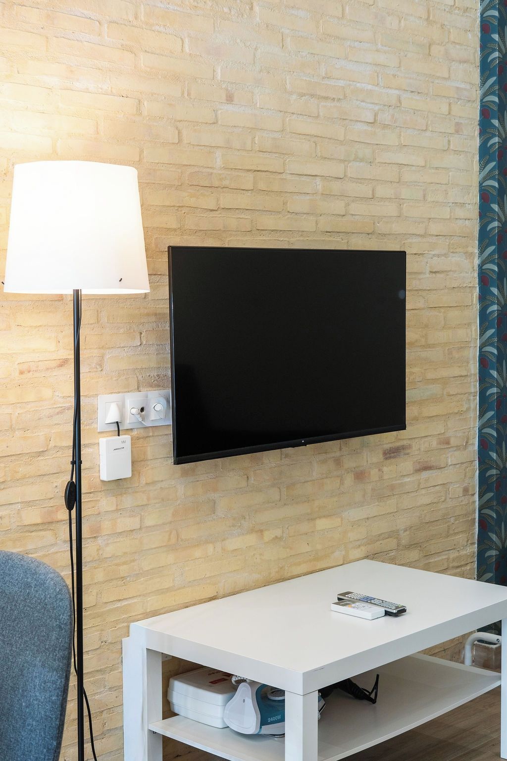 A room with a flat-screen TV mounted on a brick wall, a white table with a remote control, and a lamp on a floor stand. - VLC HOST - Fray Pedro E (Valencia)