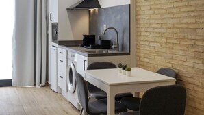 Une cuisine compacte au design moderne, dotée d’un four encastré, d’une machine à café et d’un évier. On y trouve une table à manger avec des chaises, ainsi qu’une petite plante en pot posée sur la table.