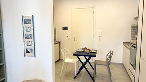 Una pequeña cocina con una mesa, sillas y una cesta. Se observa una puerta y una pared con un cuadro enmarcado.