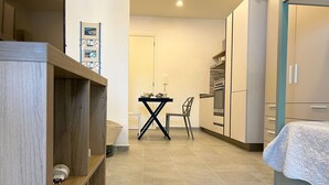 Un dormitorio pequeño con cama, una mesita con silla, y una kitchenette con armarios y microondas.