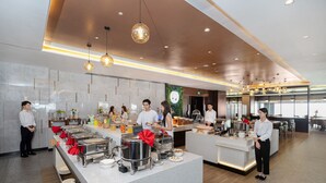 A modern hotel kitchen with buffet stations, staff serving guests, and a large window providing natural light. - JD POLMAN Hotel (Phnom Penh)