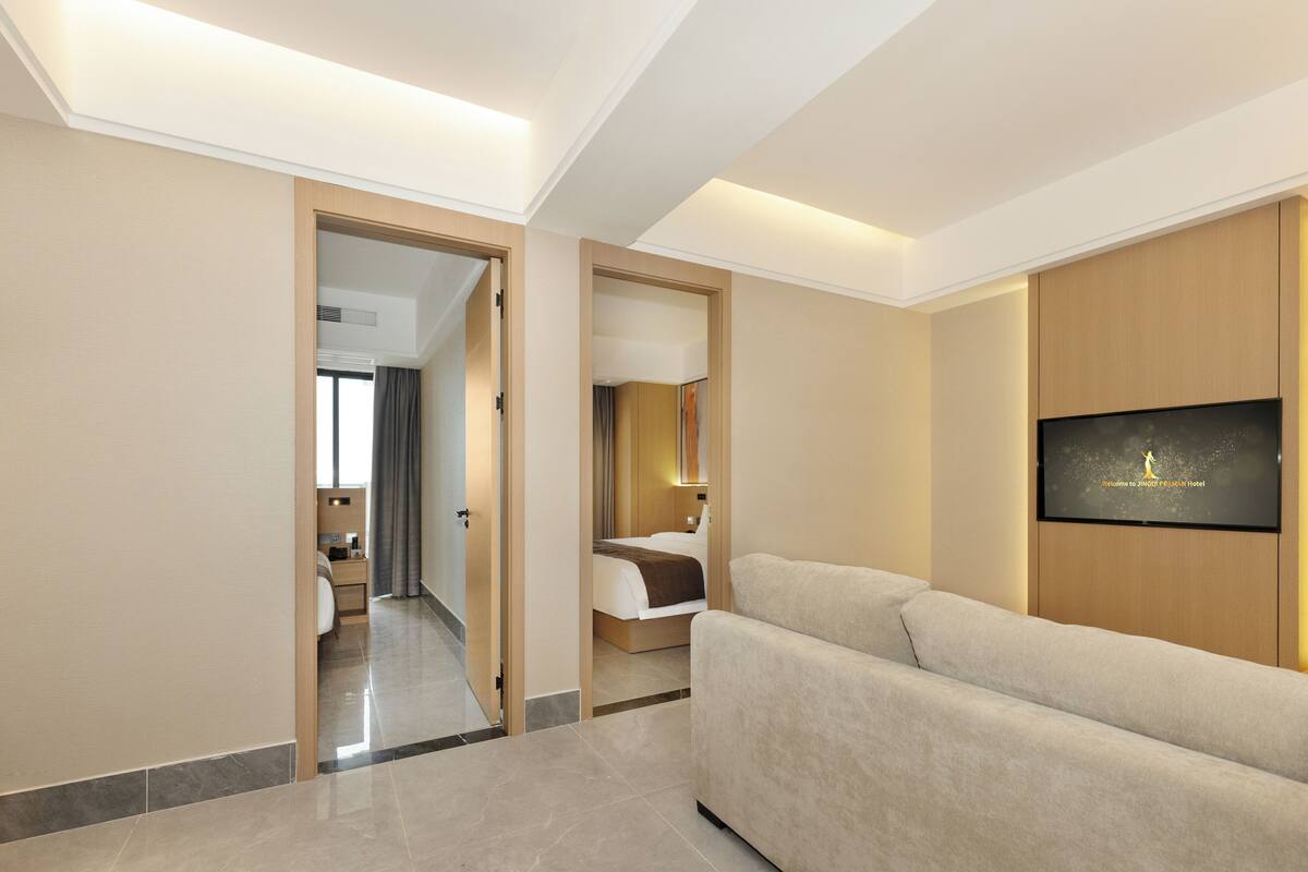 A modern hotel room with a large bed, a sofa, a TV mounted on the wall, and a hallway leading to another room.