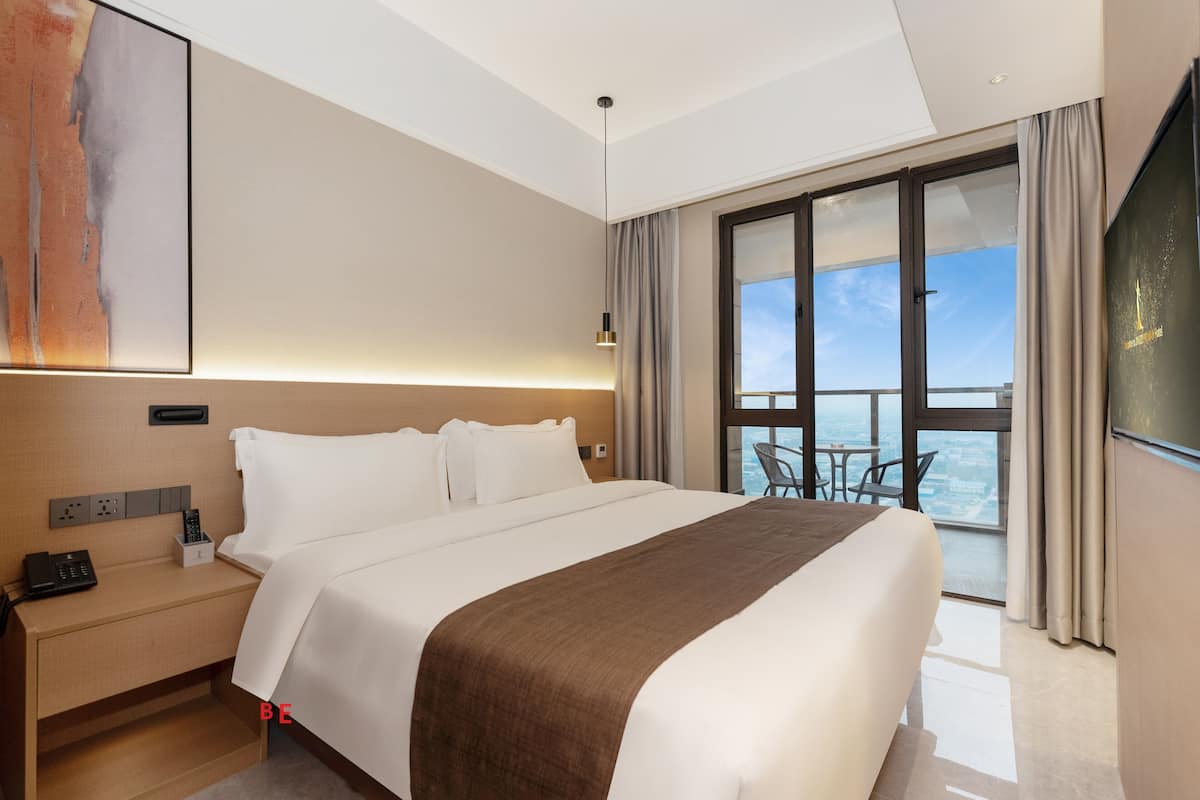 A modern hotel room with a large bed, a bedside table with a phone, a painting on the wall, and a balcony with a view.