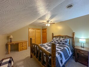 Ein Schlafzimmer mit schrägen Wänden, einem hölzernen Bettgestell, einer gesteppten Bettdecke, einem Schreibtisch mit Lampe und einem Kleiderschrank.