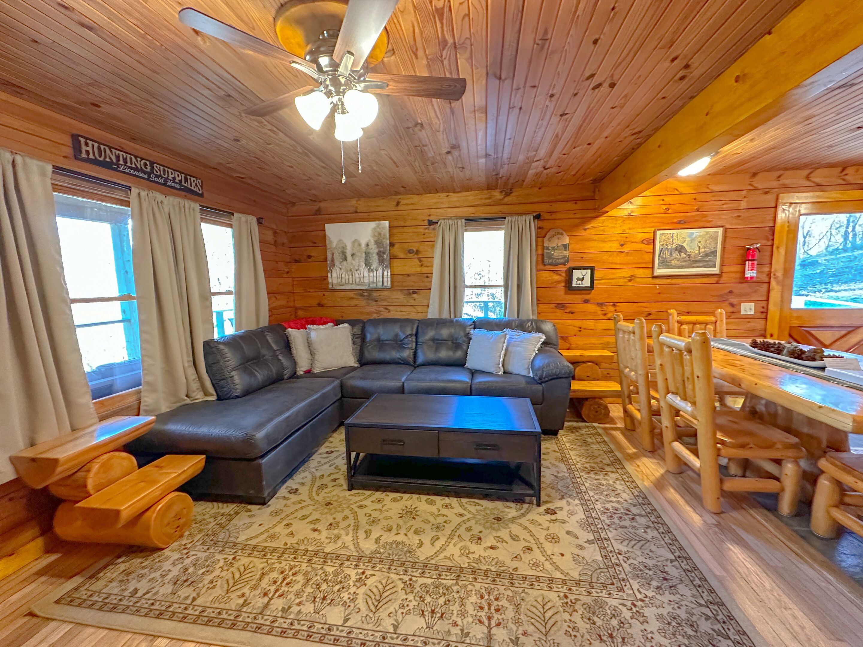 L’intérieur d’une cabane en rondins est aménagé avec un canapé d’angle, une table à manger avec des chaises, un ventilateur de plafond et un panneau indiquant où trouver du matériel de chasse.