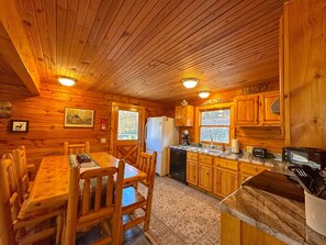 Interior de uma cabana de madeira com uma mesa de jantar, cadeiras, um refrigerador, uma cafeteira e uma janela com vista para árvores.