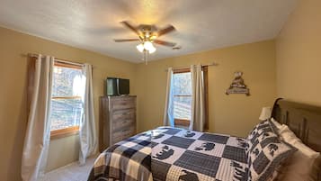 Um quarto com cama, ventoinha no teto, cômoda e janela com cortinas.