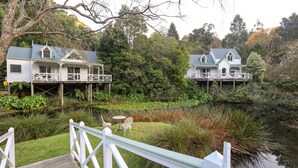 Deux maisons situées au bord de l’eau, dotées de terrasses surélevées, entourées d’une végétation luxuriante et bordées d’une étendue d’eau calme.