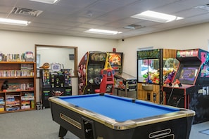 A room with a pool table, arcade games, and a bookshelf with games.