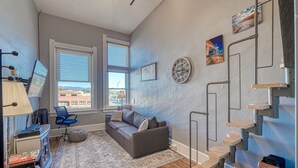 A modern living room with a gray sofa, a blue chair, a wooden staircase, and a large window offering a city view. - A Charming Loft in Downtown Salida! (Salida)