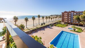 A poolside view with a beach and ocean in the background, a building with multiple floors, and a clear sky. - Beautiful apartment for 2 people with A/C, pool, WIFI, TV and terrace (Torredembarra)
