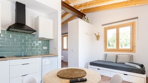 Cuina moderna amb campana extractora negra, armaris blancs i rajoles verdes. Hi ha una taula rodona amb suport tècnic trenat i un comandament a distància. Es veu un llit amb matalàs i almoinades gris a través d'una finestra.