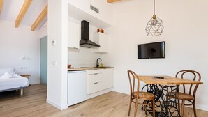 A compact kitchen with white cabinets, a wooden dining table with four chairs, a TV mounted on the wall, and a bed in the adjacent room. - Cal Tio 4 YourHouse, lovely apartment in a farmhouse with shared pool (Costitx)