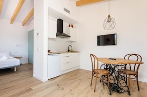 A compact kitchen with white cabinets, a wooden dining table with four chairs, a TV mounted on the wall, and a bed in the adjacent room.