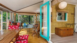 A spacious room with a wooden dining table, red cushioned benches, a wooden rocking chair, and a view of the outdoors through large windows. - Beachcombers Cottage - beachfront pet retreat (Suffolk Park)