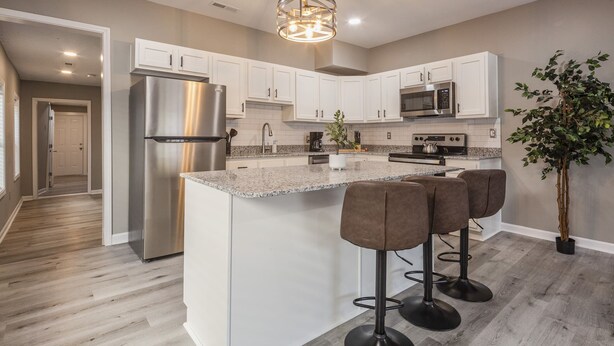 A modern kitchen with white cabinets, stainless steel appliances, a granite countertop, and a bar area with stools. - Newly renovated 4BR CozySuites (Louisville)