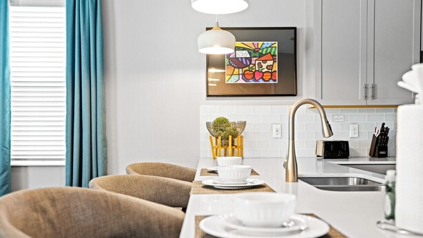 A modern kitchen with white cabinets, a stainless steel sink, and a dining area with a wooden table and chairs. - New - Sleeps 21 - Champions Gate Hyperdrive House (Kissimmee)