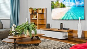 A modern living room with a large flat-screen TV, wooden shelves with plants, a coffee table with a fern, and a patterned rug. - New - Sleeps 21 - Champions Gate Hyperdrive House (Kissimmee)