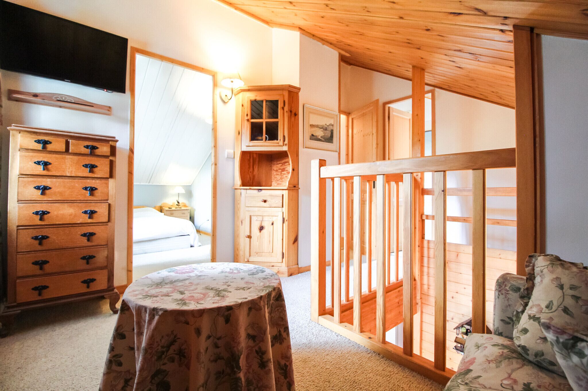 Een kamer met een houten dressoir, een bed, een tafel met een bloemrijk tafelkleed en een houten leuning.