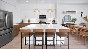 A modern kitchen with white cabinets, stainless steel appliances, a large island with bar stools, pendant lighting, and a view of the outside through a window. - Private pool & hot tub - PGA West retreat (La Quinta)