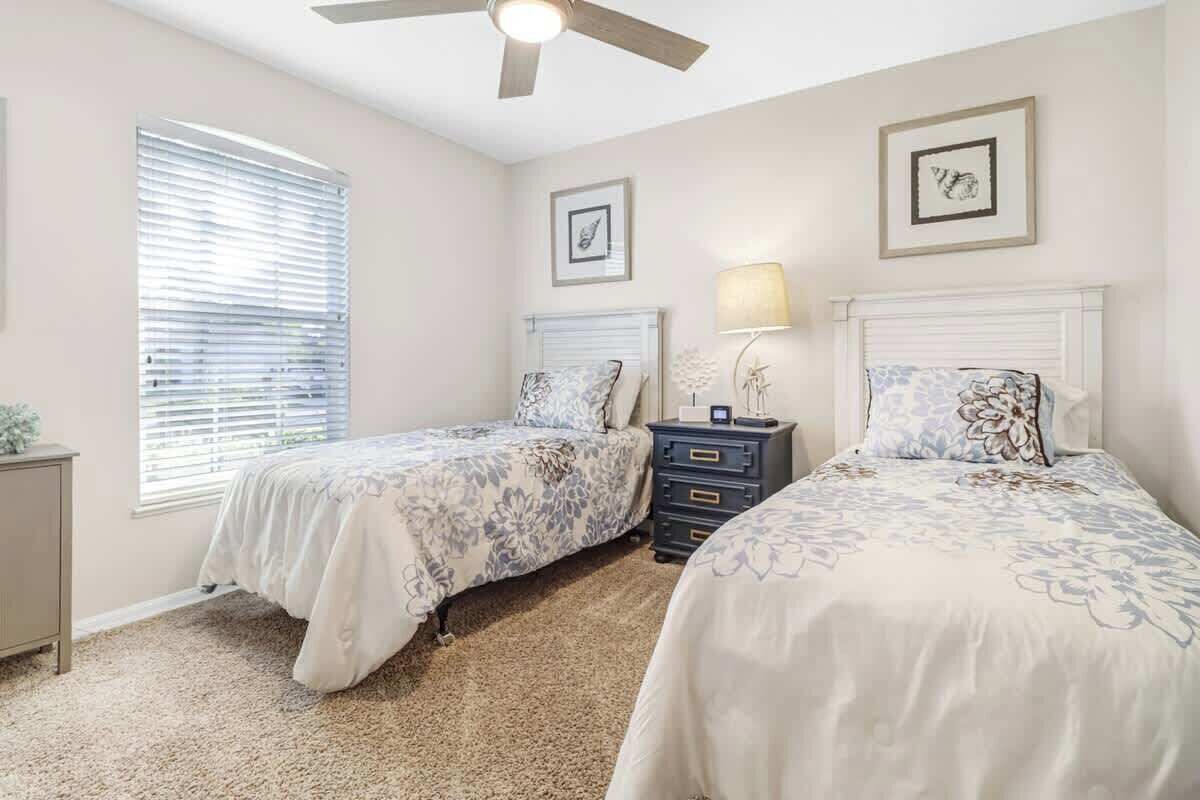 A bedroom with two beds, a ceiling fan, a nightstand with a lamp, and framed pictures on the wall.