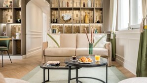 Een moderne woonkamer met een beige bank, een glazen salontafel met eten en drinken, en een decoratieve plank met diverse spullen.