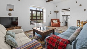 Sala de estar com sofá, poltrona e mesa de centro. Há uma televisão em um suporte, uma janela com vista para árvores e um lustre.
