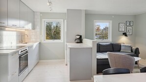 Eine moderne Küche mit weißen Schränken, einem eingebauten Backofen und einer Kaffeemaschine auf der Arbeitsplatte. Zwei Fenster bieten Blick auf Bäume und eine Straße.