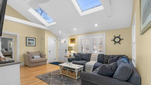 Ein Wohnzimmer mit zwei Sofas, einem Couchtisch und einem Fernseher. Im Decken befinden sich Dachfenster, eine Tür und ein Fenster mit Rollläden.