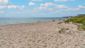 Ein sandiger Strand mit ein paar Personen und Liegestühlen, ein Rasenbereich und ein Gebäude im Hintergrund.